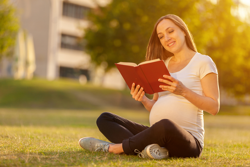 Pregnant woman is reading a book on a green field